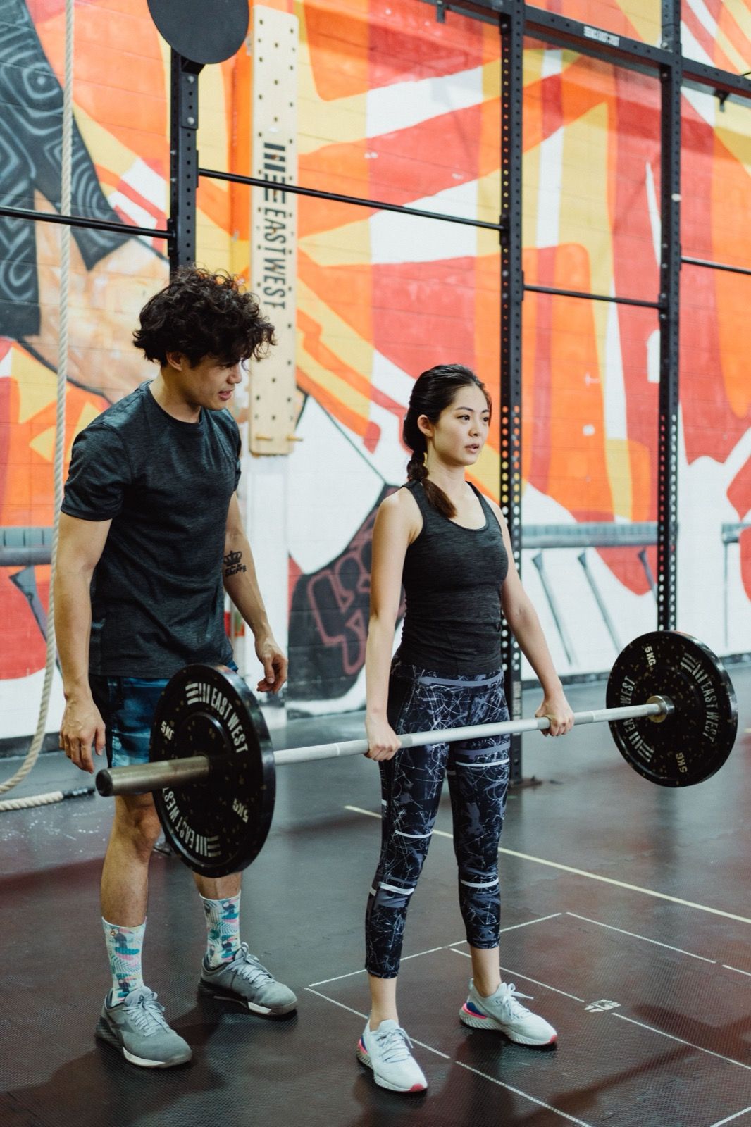 An instructor coaching a client through a barbell lift in front of a graffiti mural